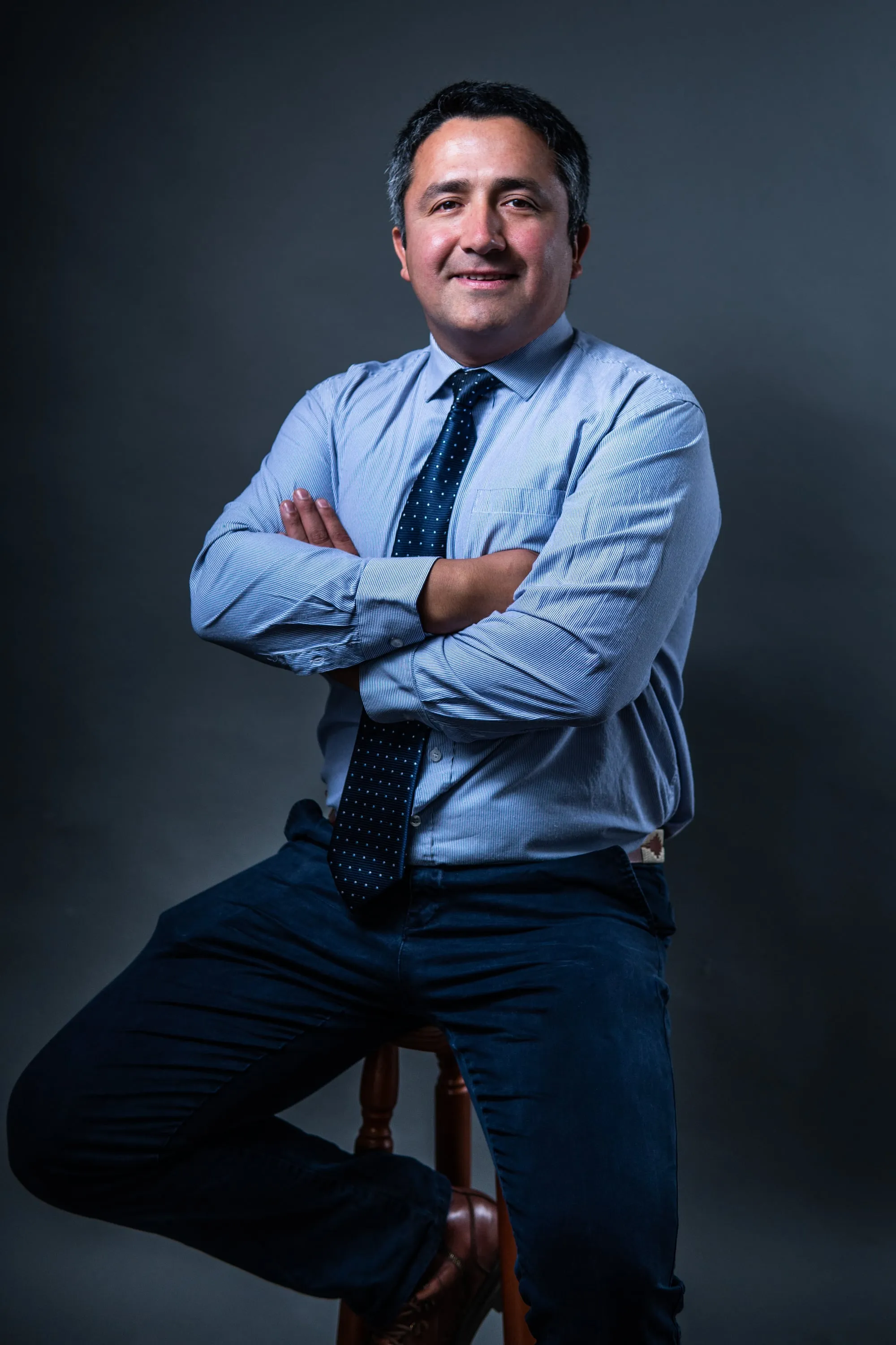 Hombre de camisa celeste y corbata, de brazos cruzados, sentado en un taburete frente a un fondo gris azulado