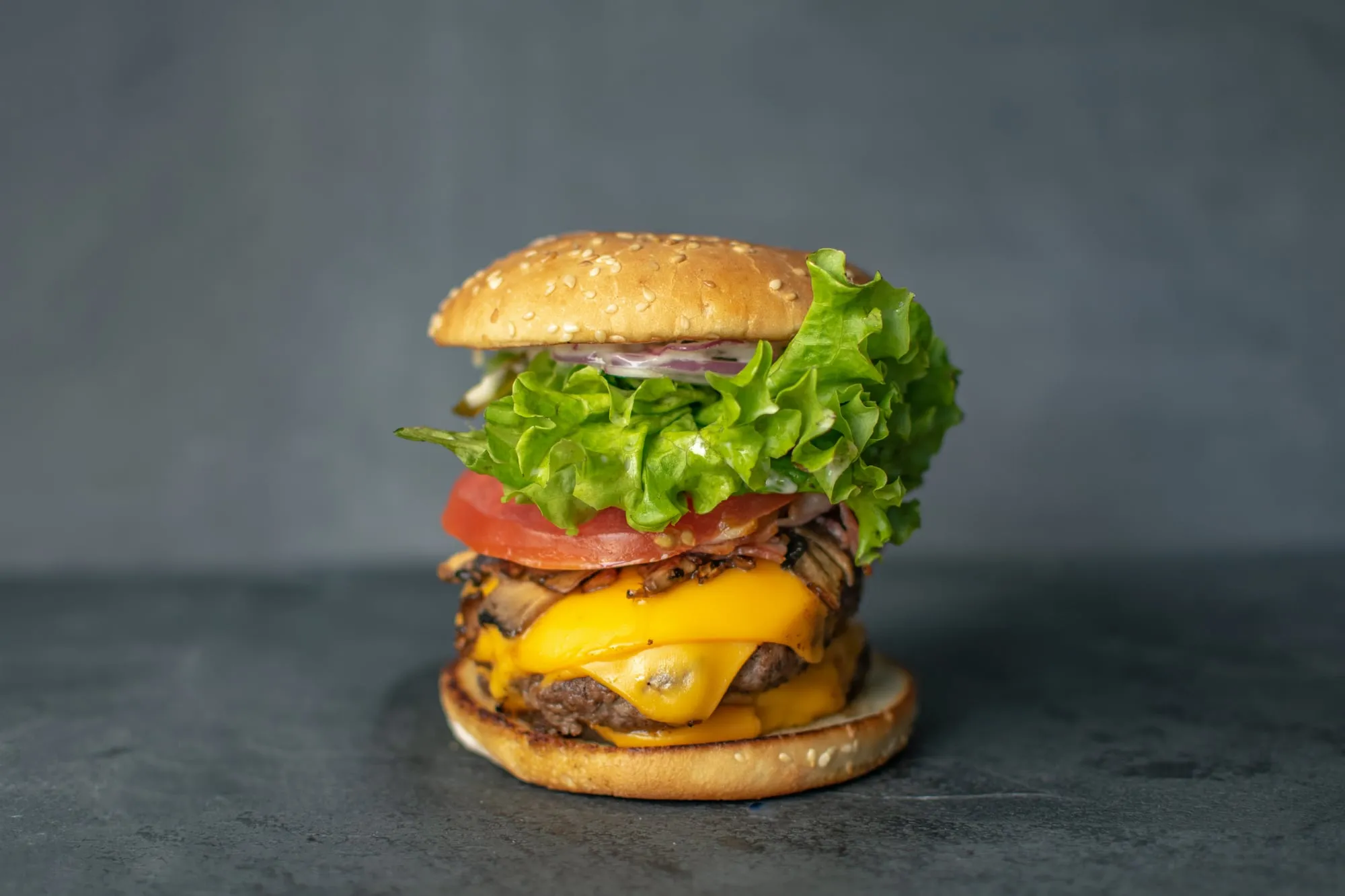 Hamburguesa alta con queso cheddar, tocino, tomate y lechuga sobre una superficie de piedra gris