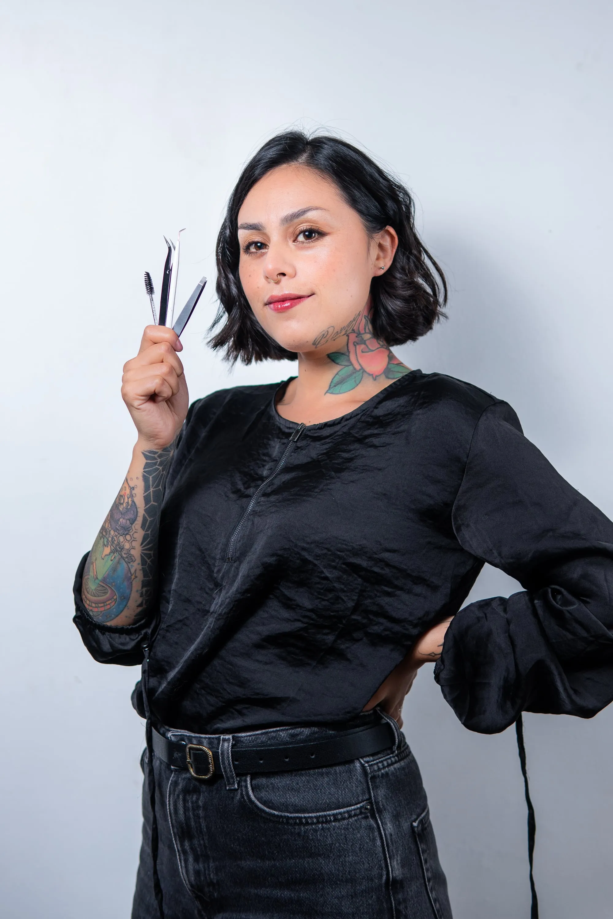 Mujer tatuada de blusa negra sostiene sus herramientas de trabajo junto al rostro, sobre un fondo claro