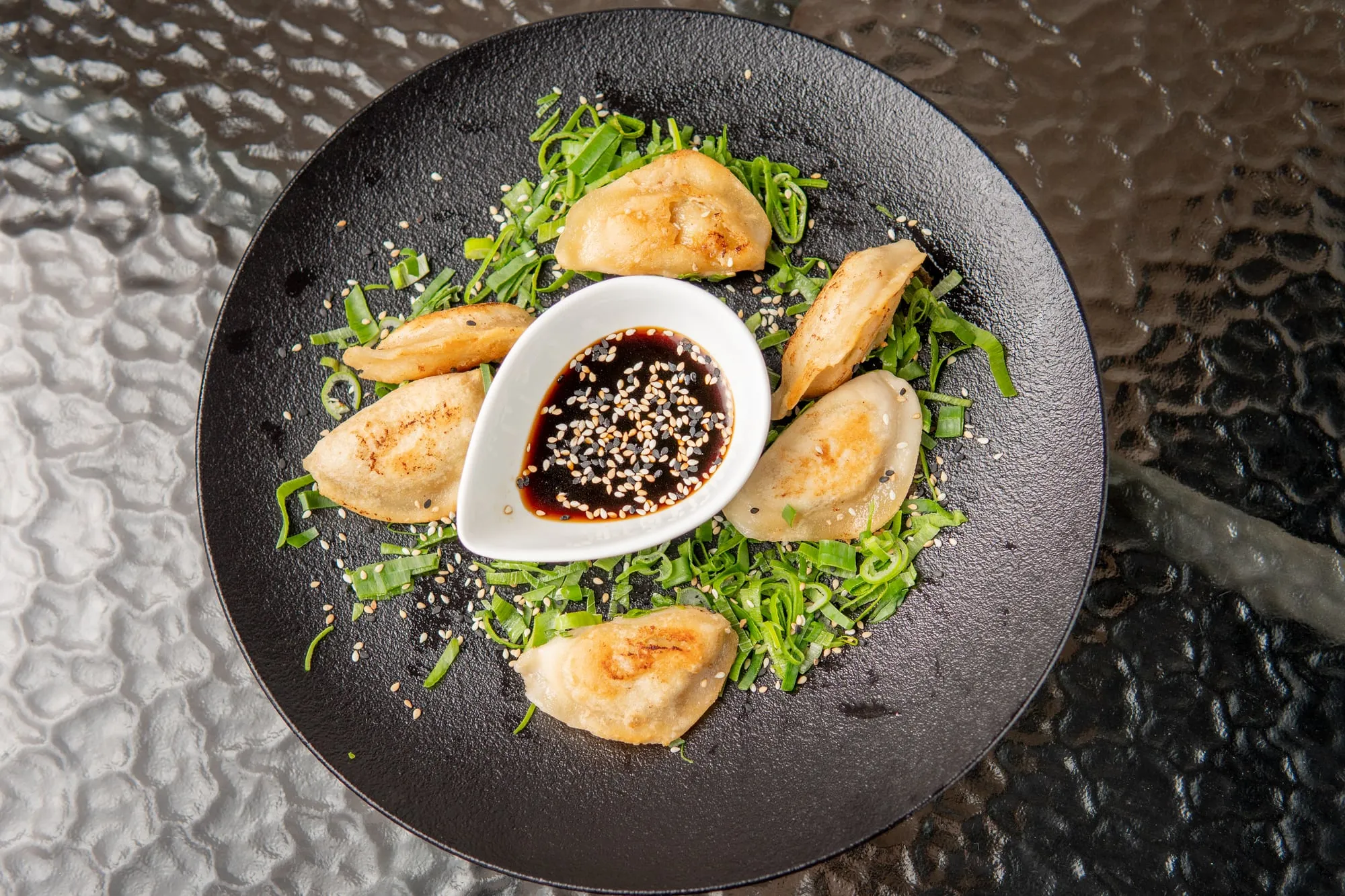Gyozas doradas alrededor de un pocillo de salsa de soya con sésamo, vistas desde arriba en un plato negro