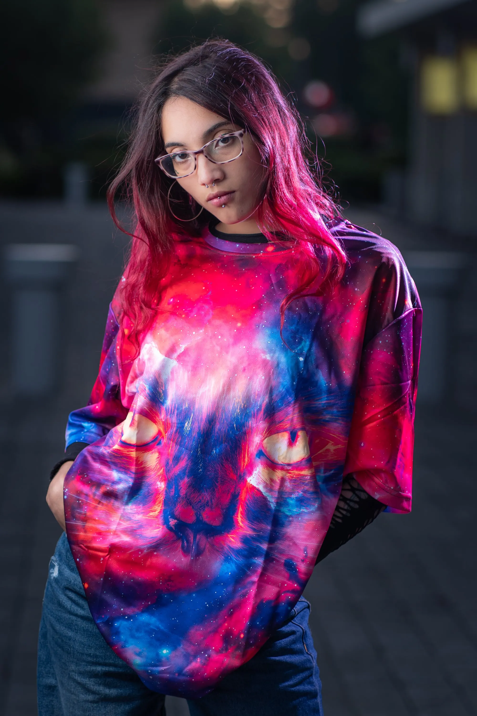 Joven de pelo rosado y lentes con polera de estampado galáctico, en la calle al anochecer