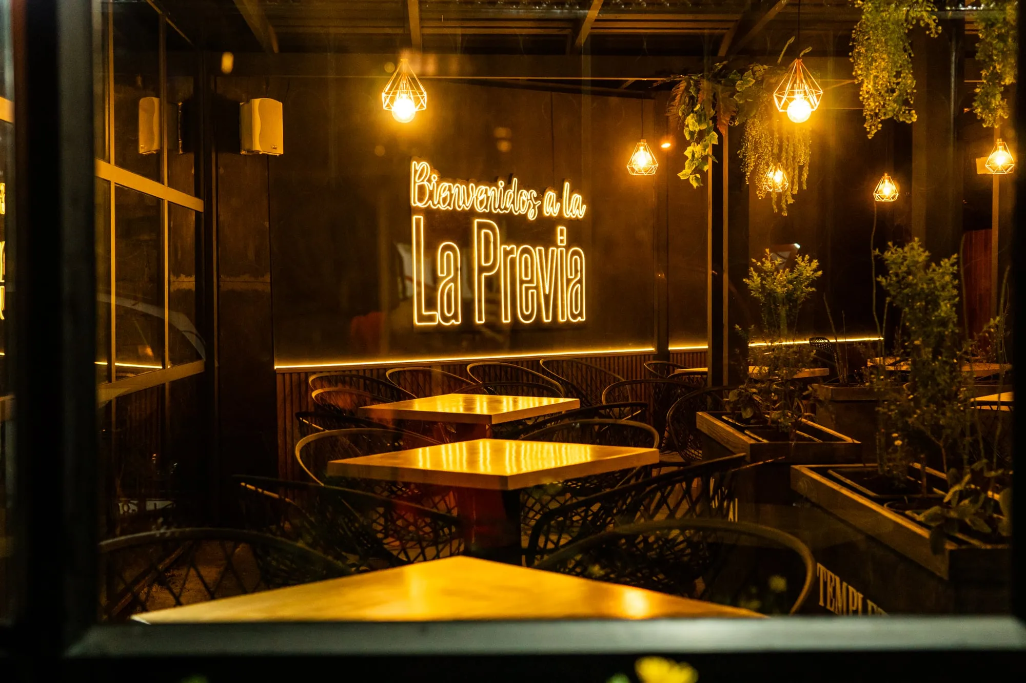 Terraza de un restaurante de noche, con mesas de madera, plantas colgantes y un letrero de neón de bienvenida